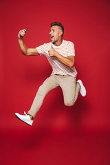 Poster - Full length photo of caucasian man in striped t-shirt jumping and taking selfie on smartphone, isolated over red background