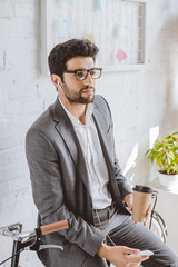 Wall Mural - businessman listening music with smartphone, holding coffee in paper cup and leaning on bike in office