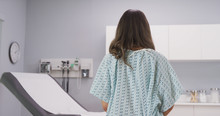 Back View Of Hospital Patient Facing Medical Equipment Inside Hospital Room. Close Up Portrait Of Female Patient Standing Indoors Medical Clinic Wearing Gown