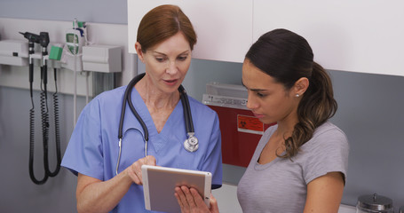 Portrait of middle aged female nurse using tablet computer to show young latina patient her medical test results. Close up of nurse explaining to young hispanic patient her health condition