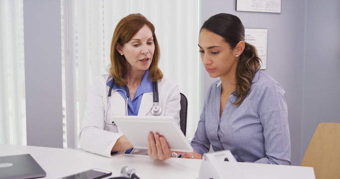 Wall Mural -  - Portrait of beautiful latina patient consulting with doctor test results on portable tablet. Charming mid aged doctor reviewing medical results on high tech device with young patient