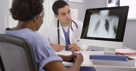 Wall Mural - Old African patient paying doctor visit to review results from x-rays of lungs. Young Hispanic radiologist showing senior African woman an x ray of her lungs on computer