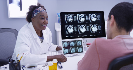 Wall Mural - Old black doctor explaining side effects of a concussion to young male patient. Senior medical physician showing patient ct-scan of brain and cranium