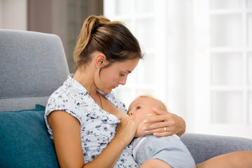 Canvas Print - Young mother, sitting at home in sunny living room, cuddling with her toddler baby boy, breastfeeding him