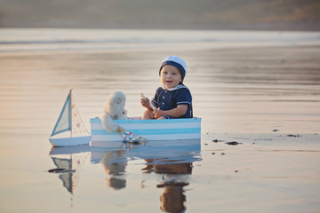 Canvas Print - Cute baby child, sweet boy, playing with boat, teddy bear and fishes on sunset at the edge of the ocean