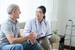 © DragonImages - Asian adult woman with clipboard sitting on sofa in house of senior man during visit and listening to patient