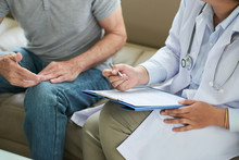 Crop Shot Of Senior Man Sitting On Sofa With Doctor During Home Visit Taking Notes On Clipboard