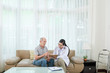 © DragonImages - Adult Asian woman showing tablet to elderly man sitting on sofa at home during visit