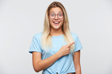 Wall Mural - Closeup of cheerful surprised young woman with blonde hair wears spectacles and blue t shirt feels happy and points to the side with finger isolated over white background