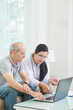 © DragonImages - Asian woman in medical gown sitting with elderly man on sofa at home and watching laptop together