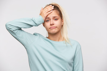 Wall Mural - Portrait of upset depressed young woman with blonde hair and ponytail wears blue sweatshirt feels ill, touching her head and having headache isolated over white background