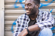 © simona - Handsome cheerful beautiful young black man african american smiling and having fun while sit and relax in the city with urban background. youthful concept for hipster style teenager boy