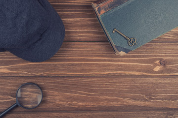 Cap of a detective, a magnifying glass and an old book with a key on the book against a worn wooden background (top view, retro style) as the detective or crime story concept