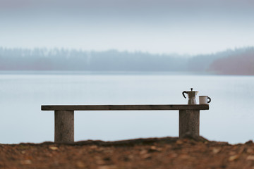 Morning. Haze. A bench on a lakeshore. On the bench there is a coffee brewer and a coffee cup.