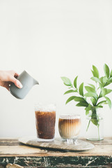 Wall Mural - Iced coffee in tall glasses with milk pouring over from pitcher by hand, white wall and green plant branches at background, copy space. Summer refreshing beverage ice coffee concept