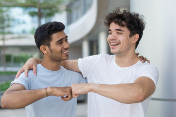 Cheerful friends making fist bump and embracing each other. Excited handsome young multiethnic men spending time together while standing at modern building. Body gesture concept