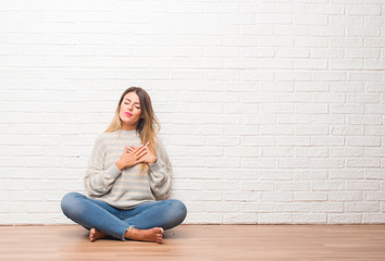 Poster - Young adult woman sitting on the floor over white brick wall at home smiling with hands on chest with closed eyes and grateful gesture on face. Health concept.