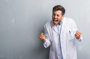 Handsome young professional man over grey grunge wall wearing white coat very happy and excited doing winner gesture with arms raised, smiling and screaming for success. Celebration concept.