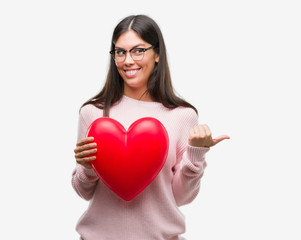 Poster - Young hispanic woman holding heart in love pointing and showing with thumb up to the side with happy face smiling