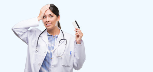 Poster - Young hispanic doctor woman holding credit card stressed with hand on head, shocked with shame and surprise face, angry and frustrated. Fear and upset for mistake.