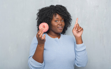 Wall Mural - Young african american woman over grey grunge wall eating pink donut surprised with an idea or question pointing finger with happy face, number one