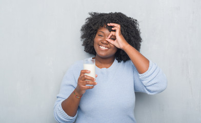 Poster - Young african american woman over grey grunge wall drinking a glass of milk with happy face smiling doing ok sign with hand on eye looking through fingers