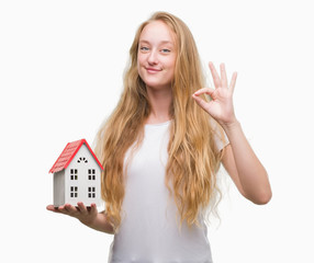 Sticker - Blonde teenager woman holding family house doing ok sign with fingers, excellent symbol