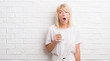© Krakenimages.com - Adult caucasian woman over white brick wall drinking glass of milk scared in shock with a surprise face, afraid and excited with fear expression