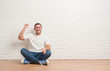 © Krakenimages.com - Young caucasian man sitting on the floor over white brick wall angry and mad raising fist frustrated and furious while shouting with anger. Rage and aggressive concept.