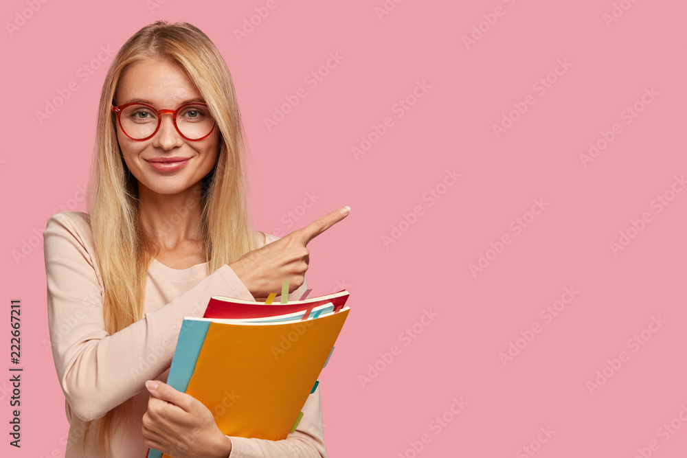 Studio shot of good looking Caucasian teacher attracts your attention to copy space, holds books, wears round spectacles, explains material to pupils, points with index finger on pink background - obrazy, fototapety, plakaty 