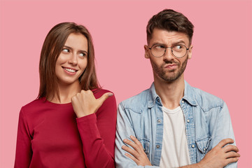 Wall Mural - Photo of cheerful young woman points with thumb at hesitant boyfriend, suggests asks him about what happened, stand together against pink studio wall. Look at my displeased uncertain best friend