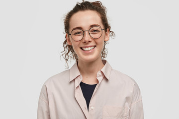 Happy young journalist has broad smile, being in good mood, has pleasant talk with people, has curly dark hair, dressed in formal fashionable shirt, stands against white background. People and fun