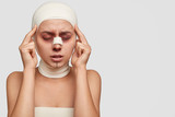 Fototapeta  - Stressful sick woman keeps index fingers on temples, closes eyes, feels pain after unsuccessful surgery operation, stands against white background with copy space for your promotion or slogan