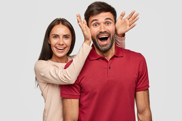 Canvas Print - Overjoyed funny girlfriend and boyfriend foolish together, have happy expressions, pray fools indoor. Positive European woman makes ears to guy with both palms, have fun during weekend at home