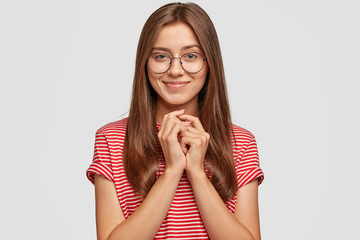 Studio shot of Caucasian woman with dark hair, appealing appearance, keeps hands together, looks with mysterious expression, anticipates for something pleasant, isolated on white studio wall