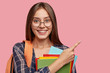 © wayhome.studio  - Horizontal shot of pleasant looking cheerful European girl points with index finger at blank copy space, advertises free space for your advertisement, carries textbooks and vocabularies, isolated