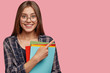 © wayhome.studio  - Studio shot of good looking young woman with dark hair, dressed in checkered shirt, points with index finger at copy space, holds books, advertises her university or college poses over pink background