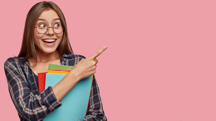 Photo of good looking woman with dark hair, pleasant appearance, indicates with index finger at upper right corner, has good mood after lectures, poses over pink background with blank space.