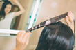 © estradaanton - Close up of hair curling device Brunette uses it for making hair style. She stands in front of mirror in bathroom. It is lightened with white light. She is concentrated.