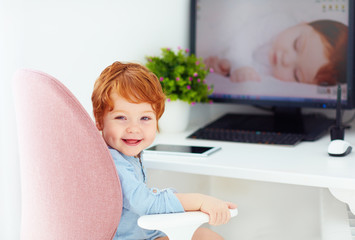 happy redhead toddler baby boy is sitting in office chair at working place