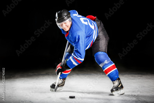 Fototapeta na szkło Young hockey player with stick and puck skating on rink in attack against dark background