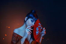 A Beautiful Geisha With Blue Long Hair And A Bang Is Looking In Soul. Red Make Up, Lips, Long Dress With Open Shoders And Deep Neckline. Holding Scarlet Fan In Hands. Japanese East Style, Art Photo