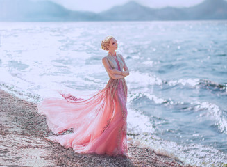 lovely slender girl in a light pink dress is walking along the sandy shore of the beautiful blue sea. glare of the sun is reflected in the waves. wonderful elegant princess elf with blond wavy hair