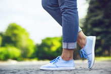 Legs Of A Girl In Jeans And Blue Sneakers On A Sidewalk Tile, A Young Woman Strolling In A Summer Park