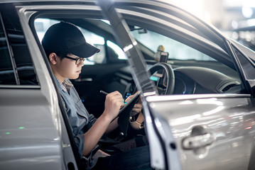 Asian auto mechanic sitting on driver seat checking the car using digital tablet in auto service garage. Mechanical maintenance engineer working in automotive industry. Automobile servicing and repair