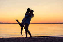 Silhouette Of Happy Couple On Beach At Sunset, Man Taking The Girl In His Arms.