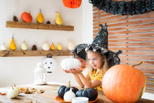 Cute Little Girl In Witch Costume Sitting Behind A Table In Halloween Theme Decorated Room, Holding Hand Painted Pumpkins And Smiling. Halloween Holiday Concept.