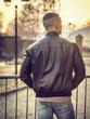 © theartofphoto - Rearview of one young man in urban setting in European city, standing, wearing black leather jacket and jeans at sunset