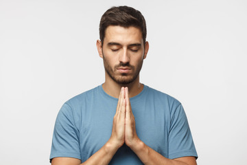 Wall Mural - Closeup of young man isolated on gray background, putting hands together as if he is praying with closed eyes