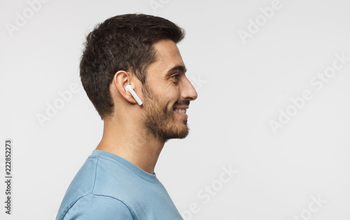 Fotografía Sideways portrait of smiling young man listening to music or radio, uses modern wireless earphones, wearing blue t-shirt